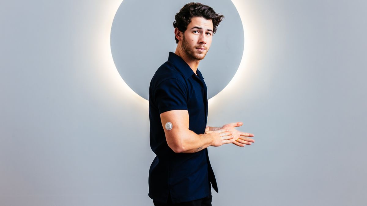 Nick Jonas poses in front of a glowing background, wearing a small, round device on his upper arm